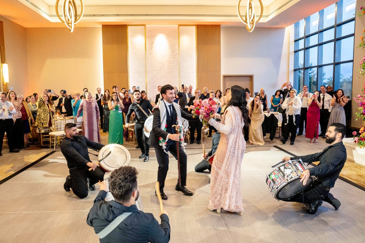 A lively wedding celebration with a couple dancing joyfully in the center. Musicians on drums perform energetically around them. Guests in elegant attire cheer and capture the moment on their phones. The hall is warmly lit with large windows in the background.