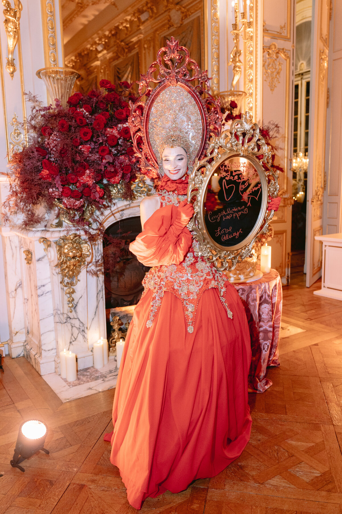 Photo was taken at Shangri La Hotel & Ritz Paris Hotel Paris, France. by photographer Matteo Coltro. A person dressed in an elaborate orange and gold gown with floral embellishments holds a decorative mirror with a congratulatory message written on it. The scene is set in a lavish, ornately decorated room with large red floral arrangements and candles on the floor. Videographers for the event were Zen Film Works.