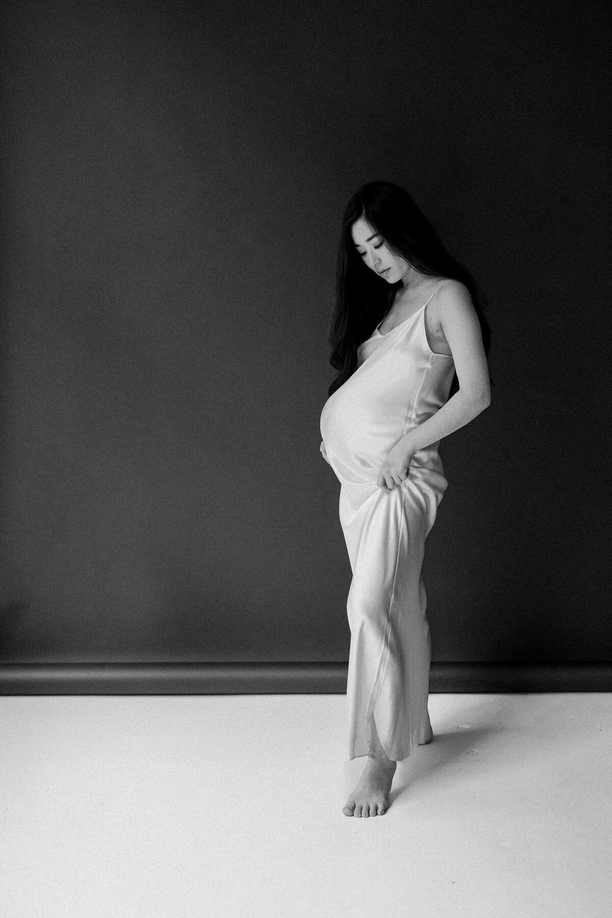Pregnant woman poses in front of black backdrop in a cream silk dress.
