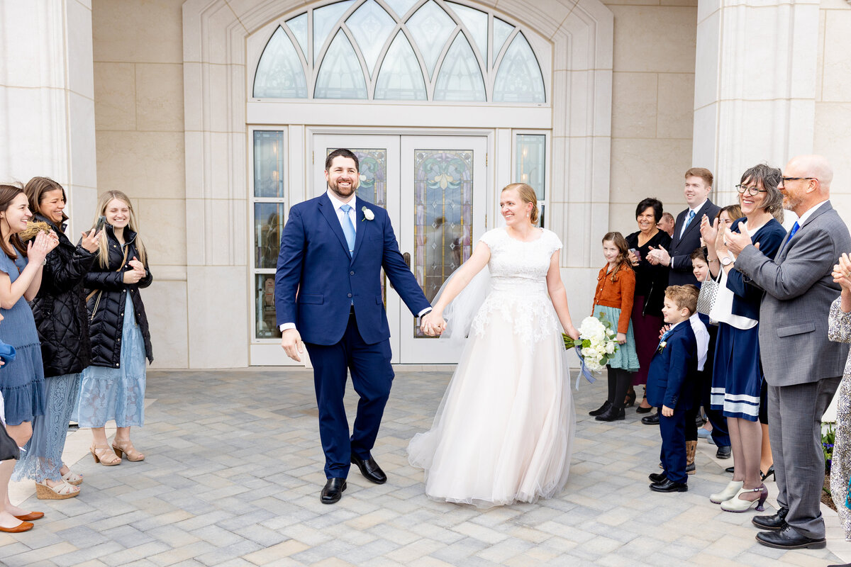The newlywed couple smiles at each other while walking hand-in-hand through a cheering crowd outside the temple doors.