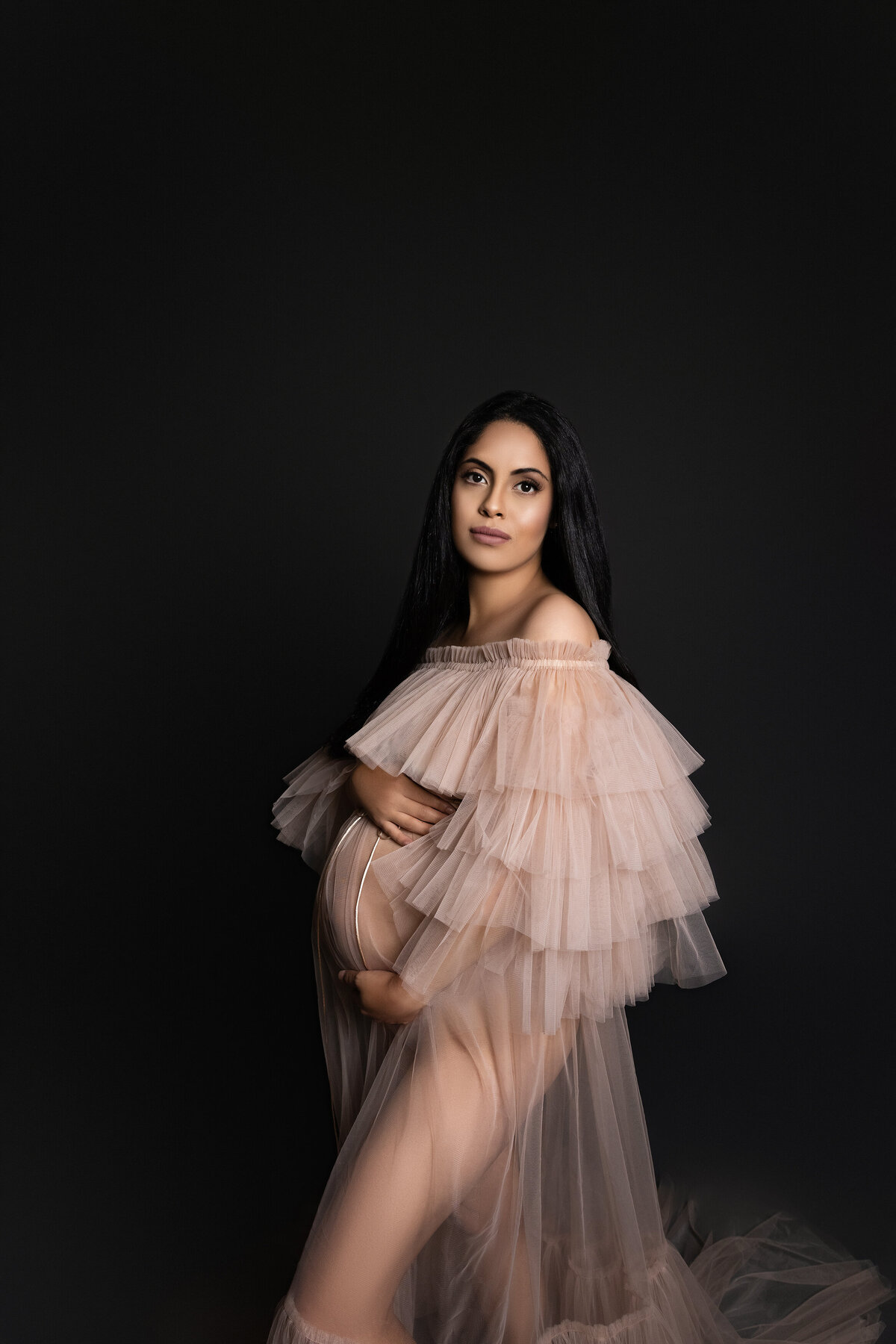 Maternity photos captured by top London, ON Maternity Photographer Amy Perrin-Ogg. Woman in tulle gown is standing side profile to the camera looking over her shoulder and holding her bump.