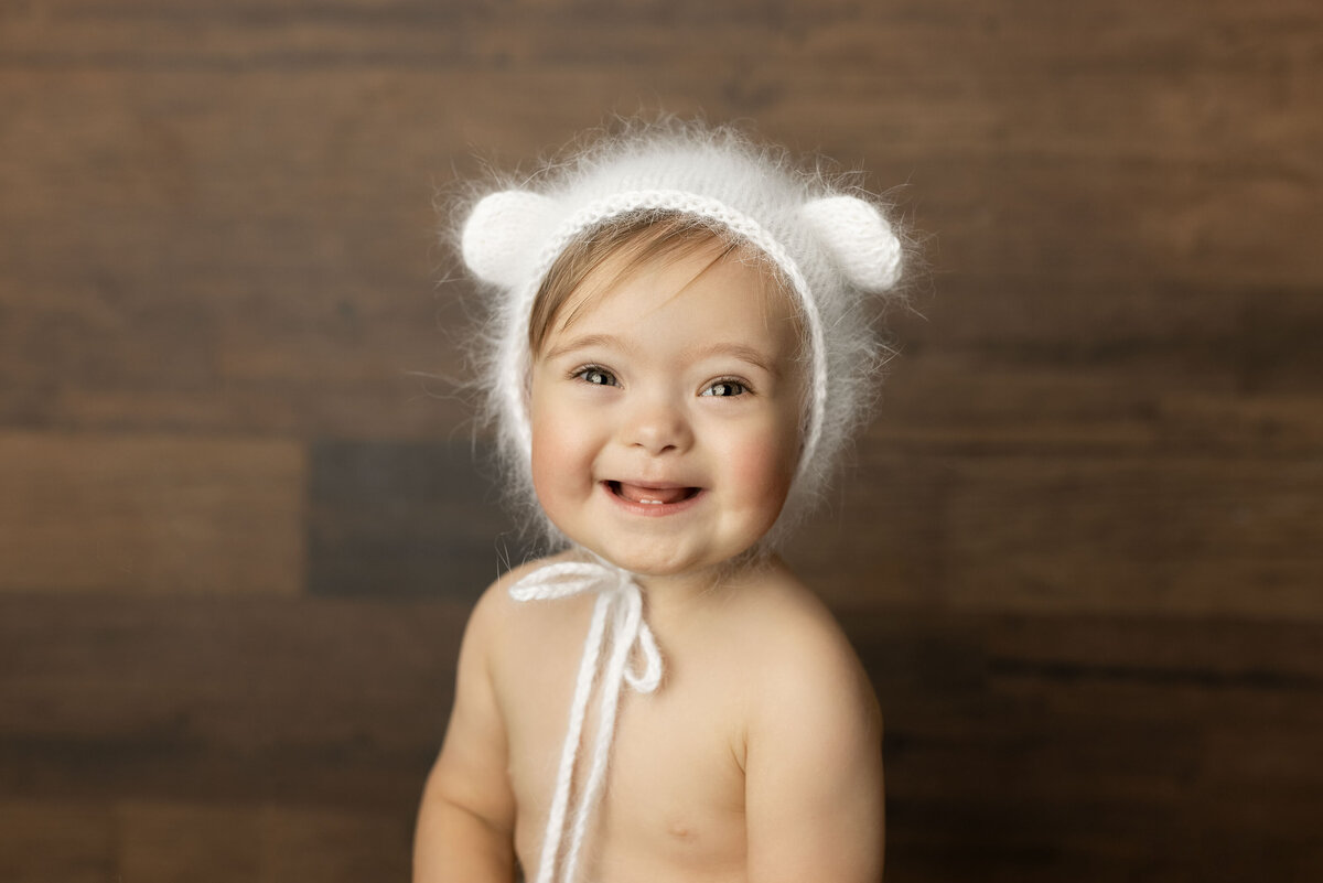 One-year-old smiling at the camera in London, ON studio wearing a white, fuzzy, teddy bear bonnet.