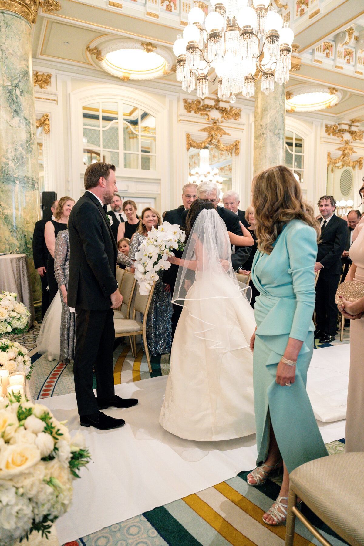 A bride in a white gown and veil walks down the aisle accompanied by a woman in a blue dress. Guests in formal attire stand in an ornately decorated room, featuring chandeliers and gold accents, as they watch the wedding ceremony.