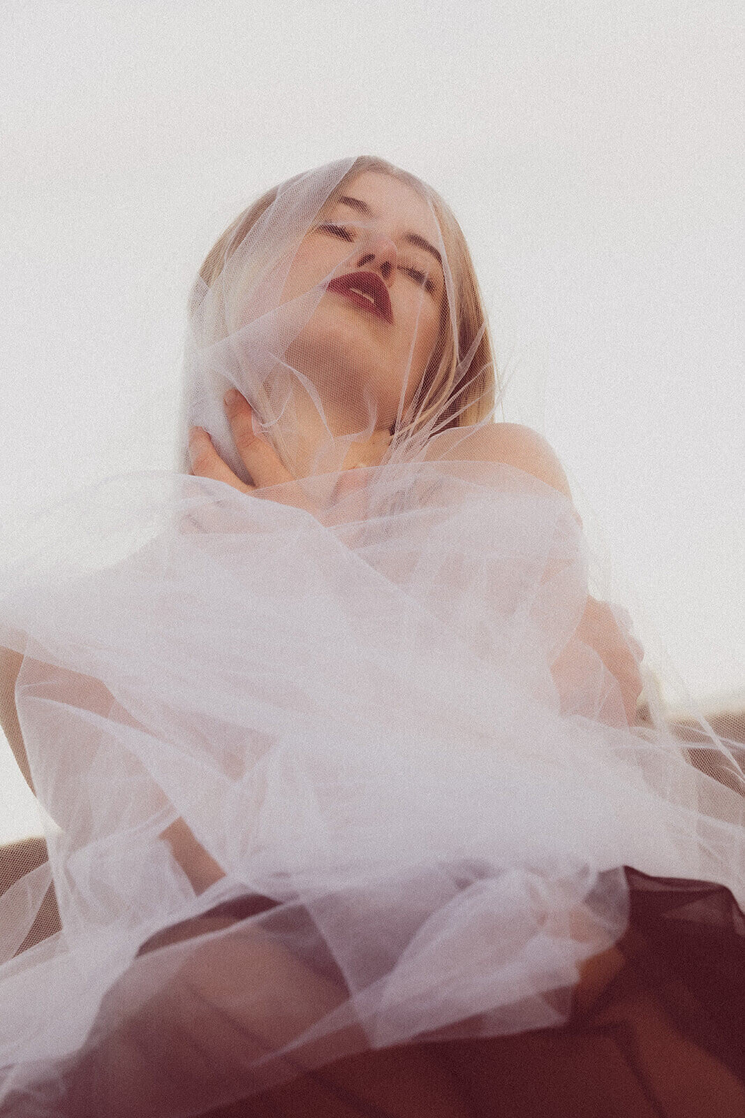 A bride with red lipstick is wrapped in tulle and embracing herself