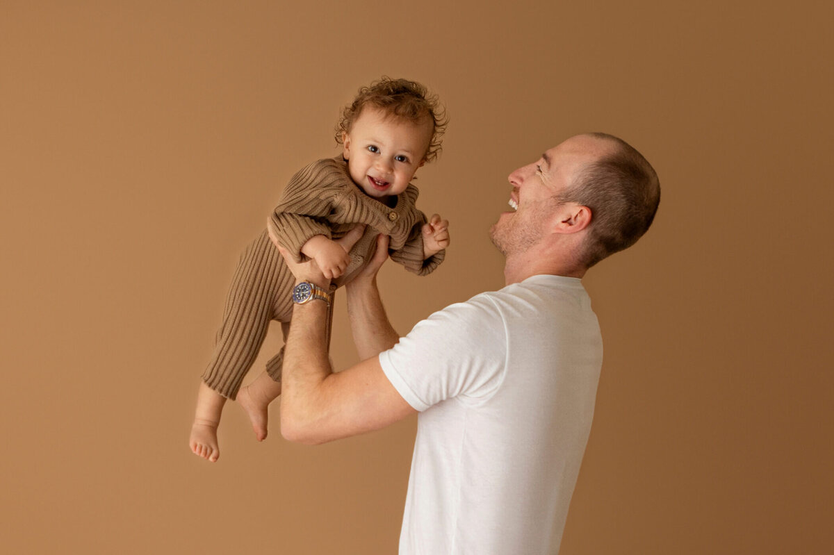 A joyful father lifts his smiling young child into the air against a warm beige background. The father, dressed in a white T-shirt, looks up at his child with a big smile. The child, wearing a cozy brown outfit, looks down at their father with a happy expression, arms outstretched. The playful interaction captures a moment of pure happiness and bonding.