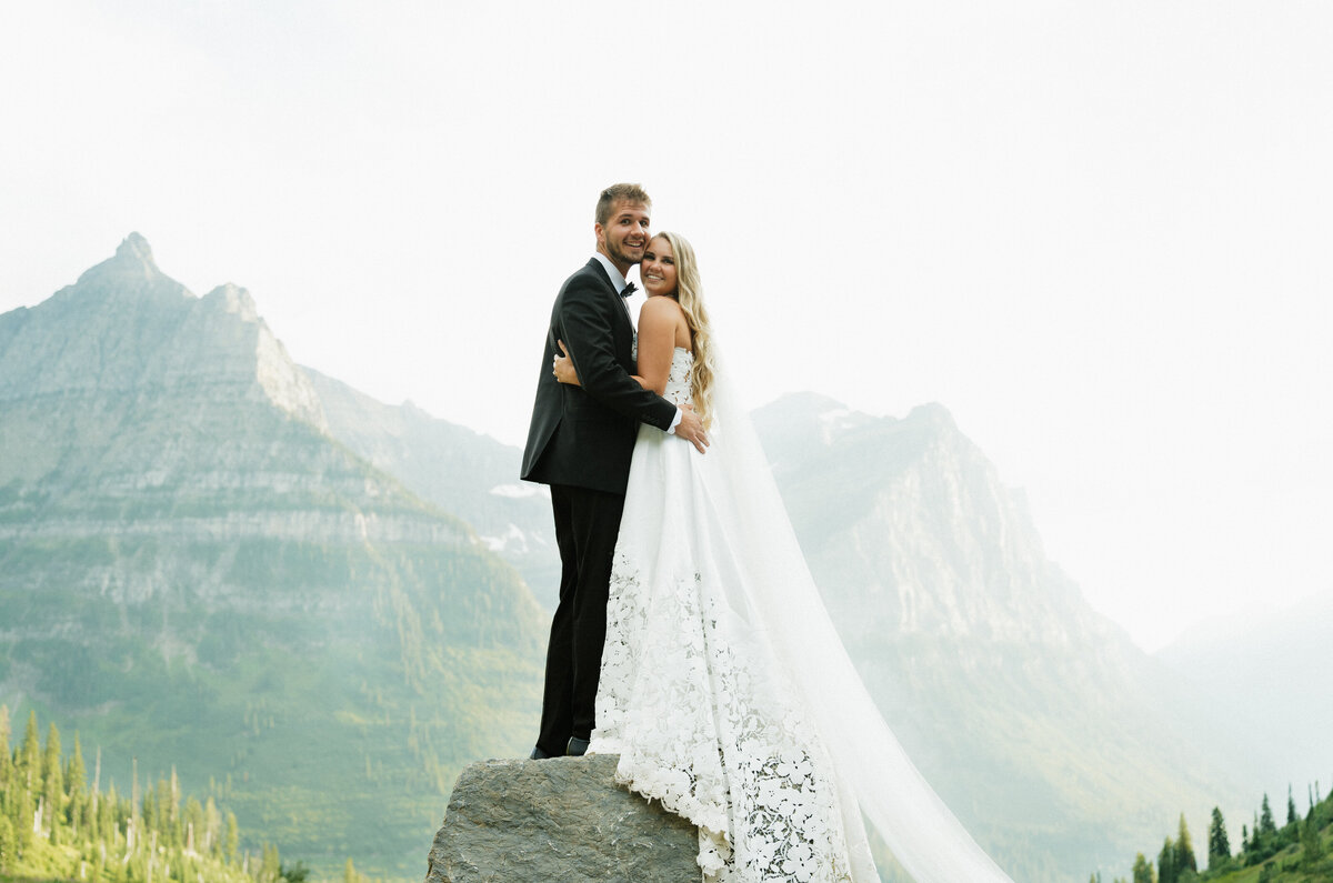 Elopement photography and wedding photography in Glacier National Park Montana. The bride and groom are facing and holding each other standing on a large rock with mountains behind.