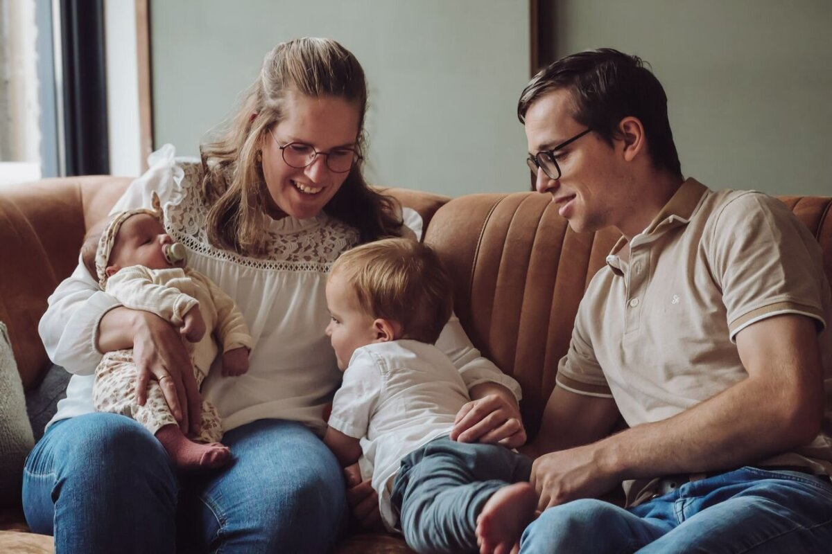 een mooi gezin zittend op een bank terwijl de moeder haar baby vasthoudt en een jonge jongen de broer kijkt naar de baby en hun vader