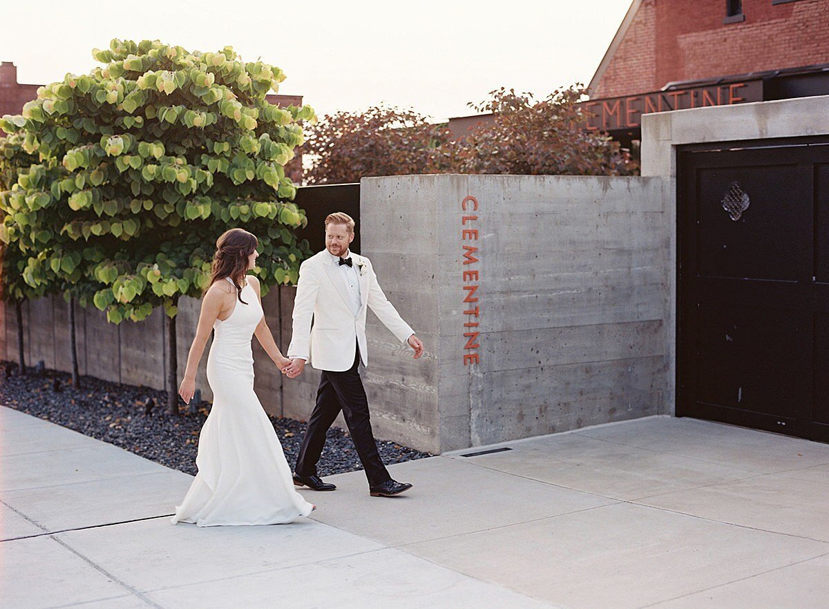 The bride, wearing a silk halter top sheath dress holds hands with the groom wearing a tuxedo with a white jacket at they walk into Clementine Hall in Nashville, TN. The gray cement wall is accented with the word Clementine written in orange.