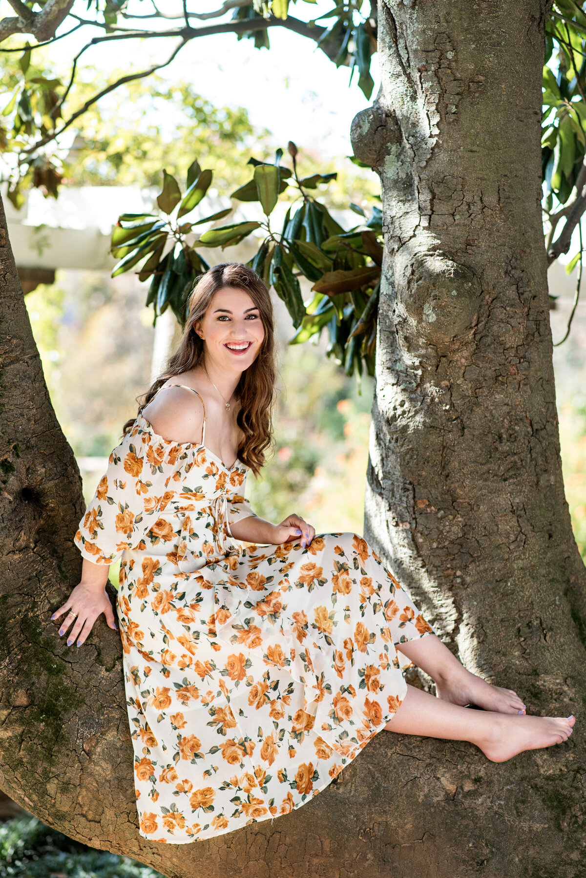 RVA senior girl sits in a magnolia tree at Maymont Park wearing floral dress duringher senior portraits.