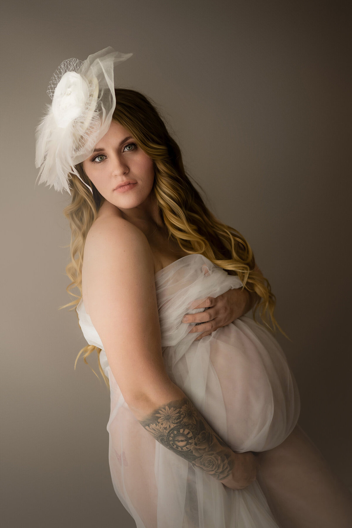 A woman stands in a studio in a tiny white hat and covering with a sheer white fabric