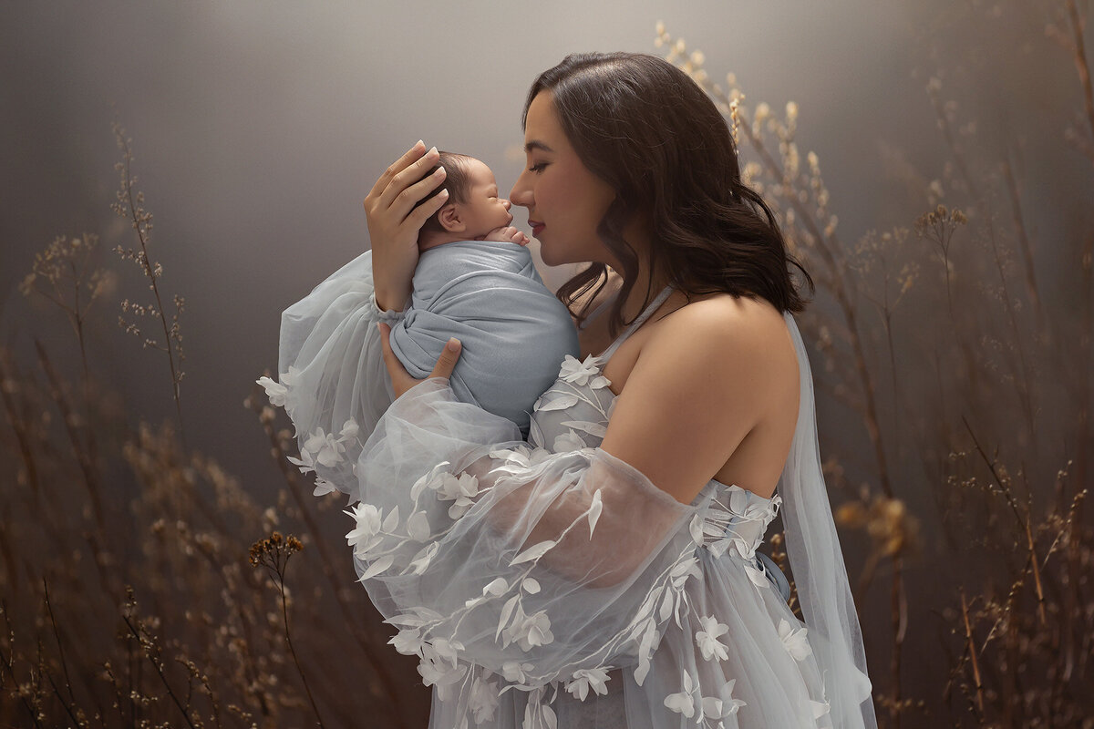 Mother in a flowing blue floral gown gently touching noses with her swaddled newborn, surrounded by soft dried florals and glowing studio light, captured by a Dallas newborn photographer.