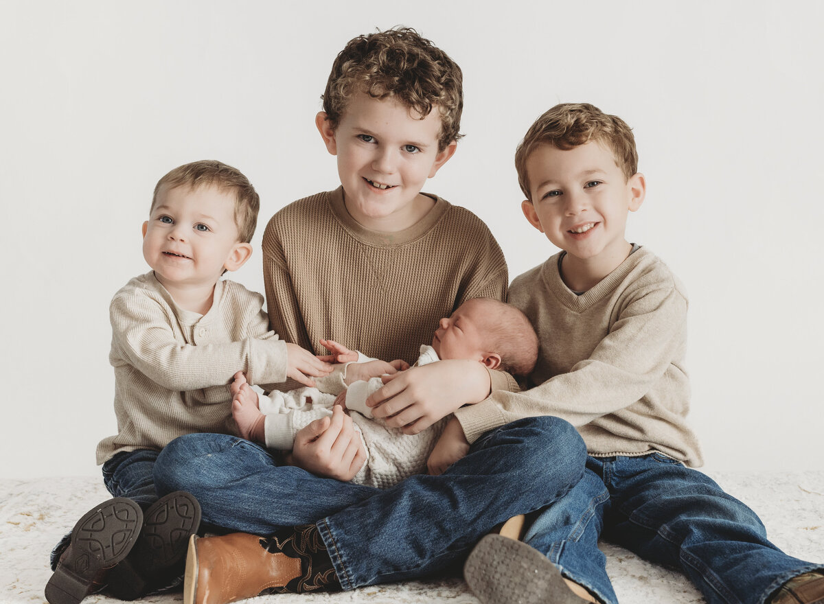 Three brothers sitting close together in a studio setting, smiling and holding their baby sibling for a sibling milestone portrait.