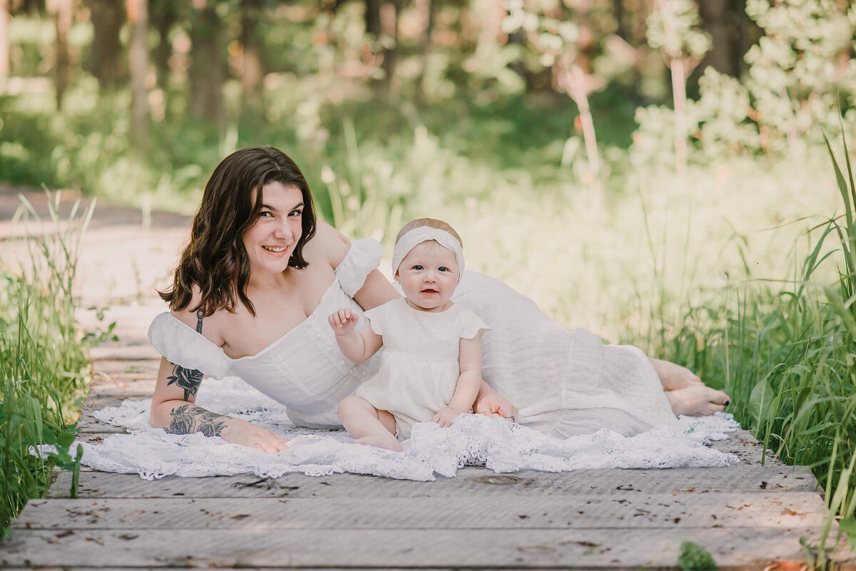A mother and infant daughter lay on a wooden bridge. Mom lays on her side and baby sits in front. They both smile at the camera. Brittany Danielle Photography