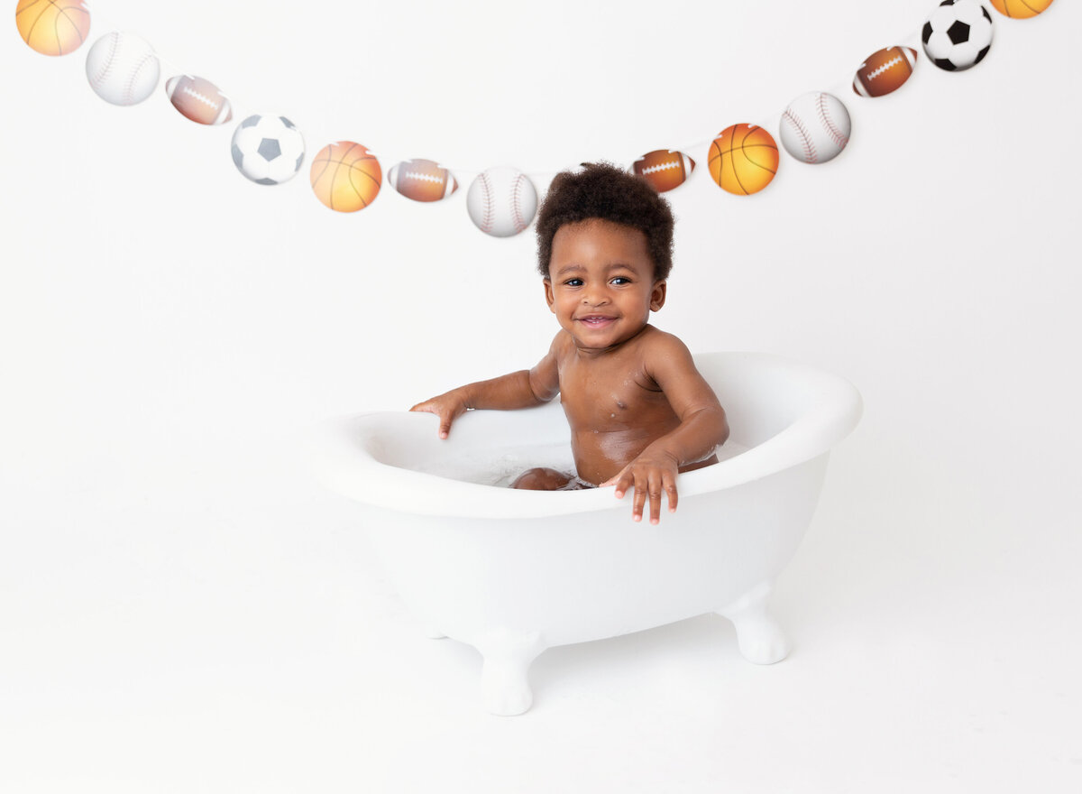 Baby boy sits in a mini bathtub cleaning up from a sports themed cake smash first birthday photoshoot.