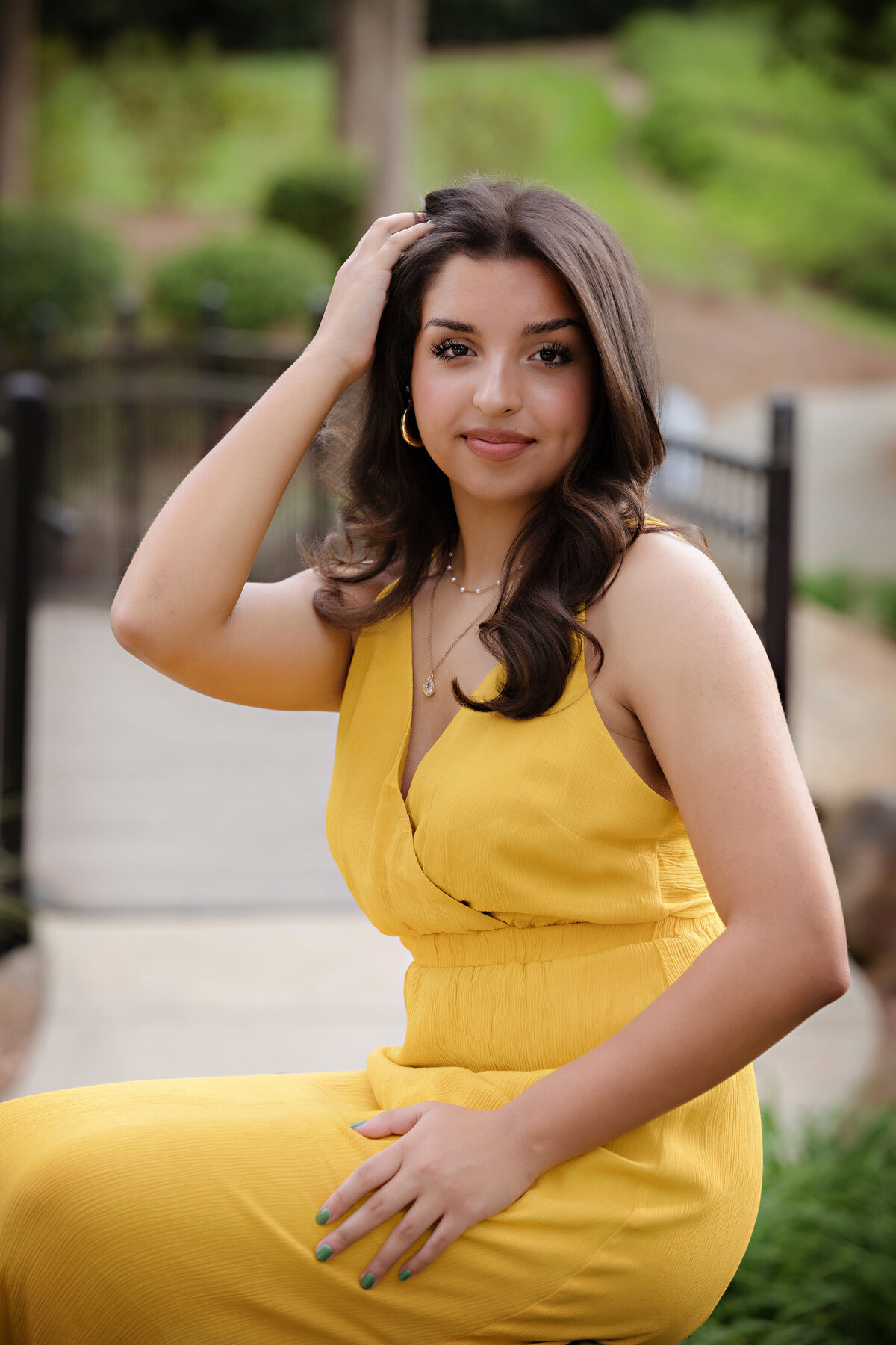 Senior portrait of teen in yellow dress sitting atop a park bench with a bridge path in the background, captured in soft spring light.