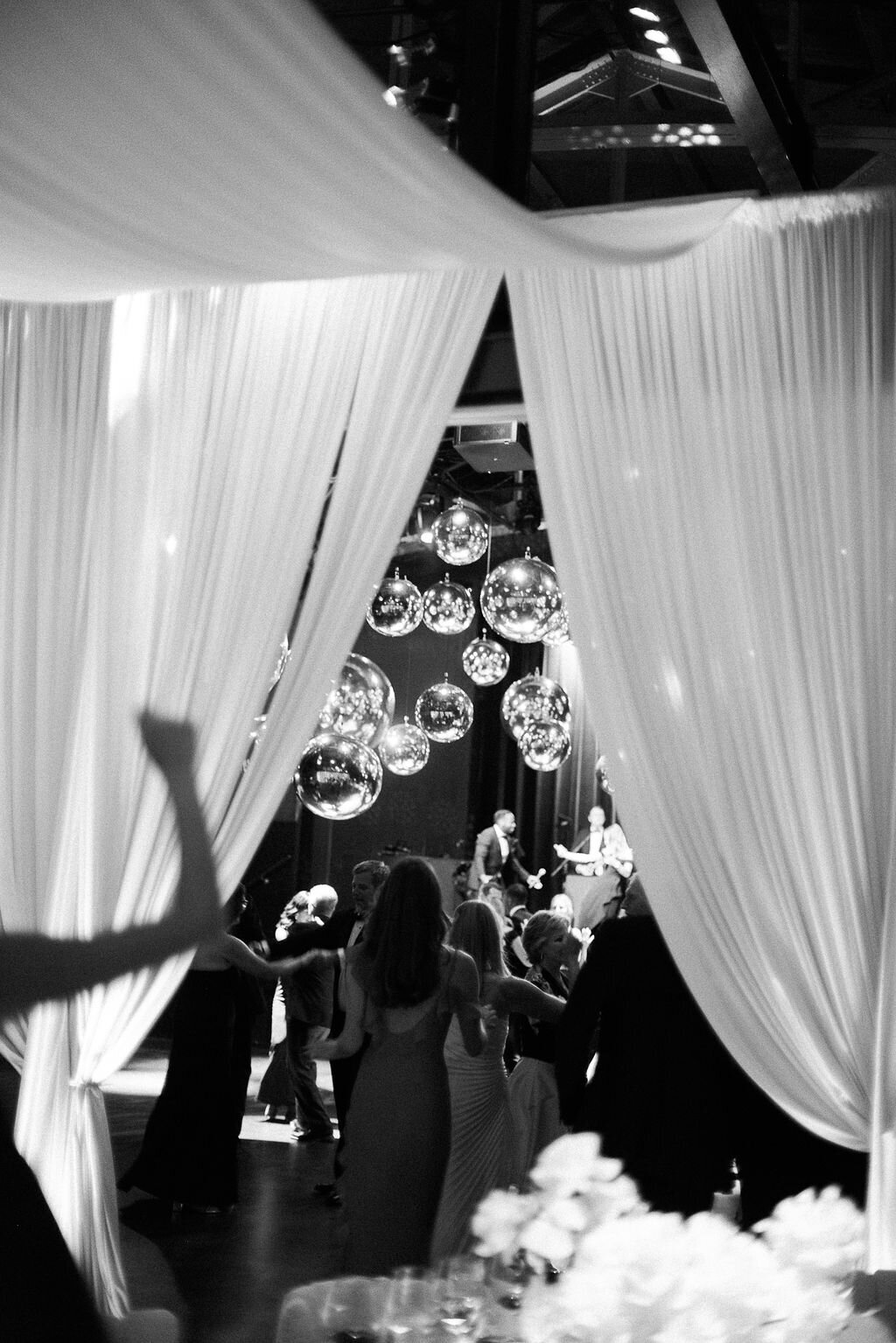 A black-and-white image of guests raising their hands in excitement as they enter a glamorous wedding reception space with hanging mirror balls.