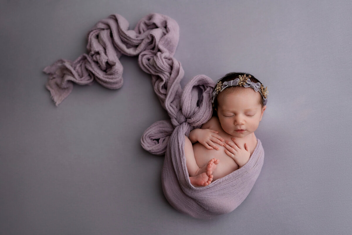 Newborn baby wrapped in a lilac swaddle, adorned with a delicate floral headband, peacefully sleeping on a grey background.