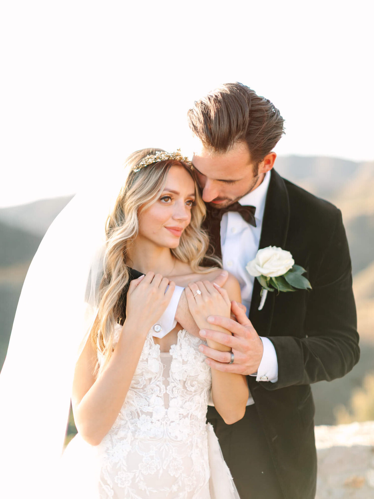 A couple embraces in wedding attire, the bride in a lace gown and veil, groom in a dark suit with a rose boutonniere.