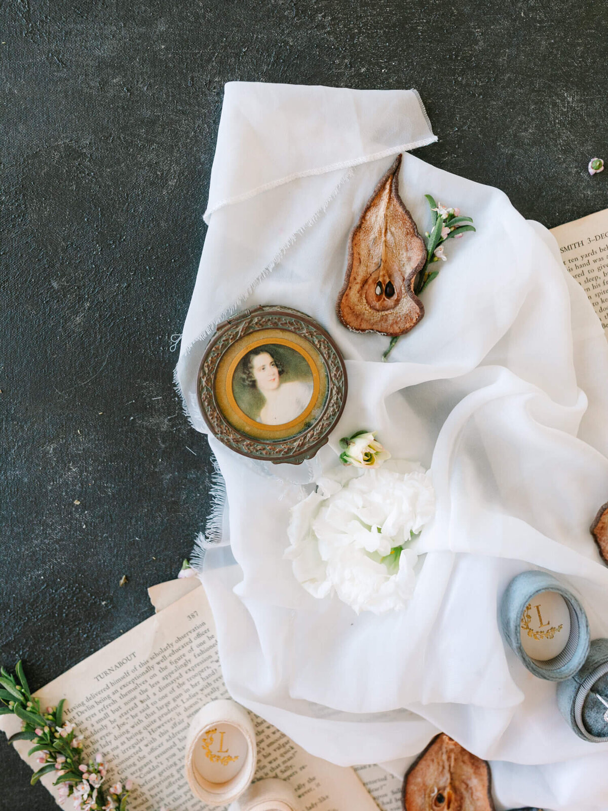 Elegant flat lay with a vintage portrait, dried pear slice, white flowers, and open book on soft white fabric.