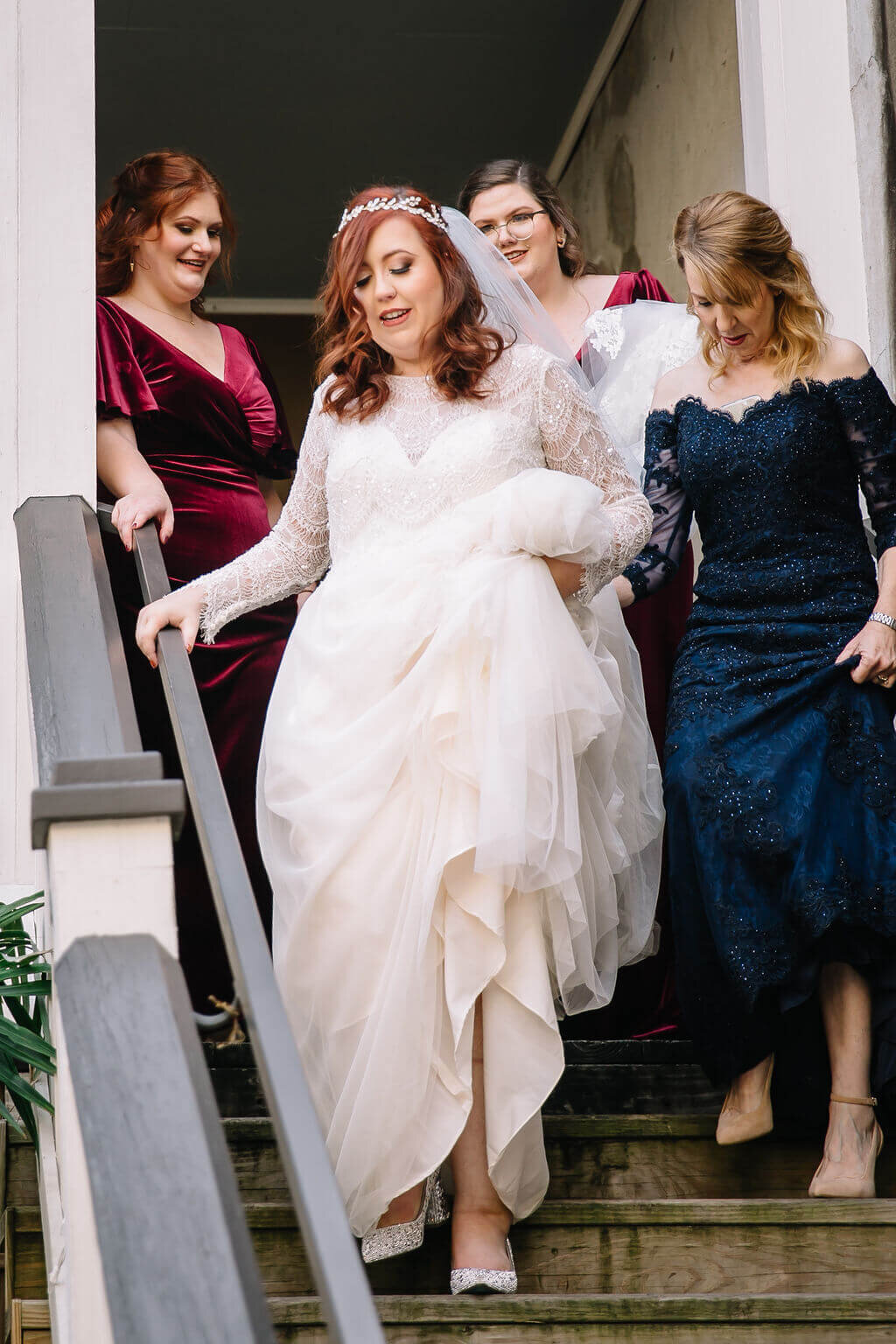 A bride in a white dress walks happily down a staircase, holding her gown, followed by bridesmaids in dark dresses. The scene is joyful and candid, with natural light streaming in from the left.