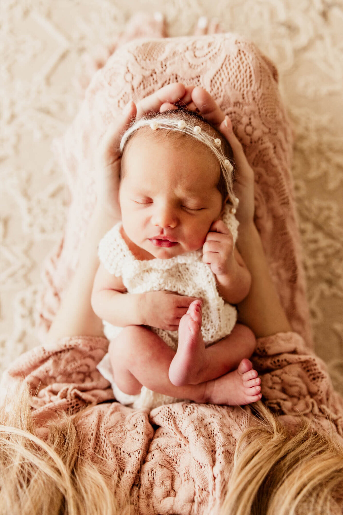 Newborn baby girl wearing a white pearl headband sleeping in her mother's hands with her legs crossed, OKC newborn photo session.