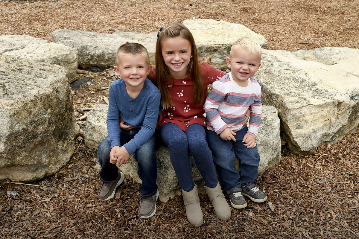 A girl and her 2 younger brothers sit on some rocks
