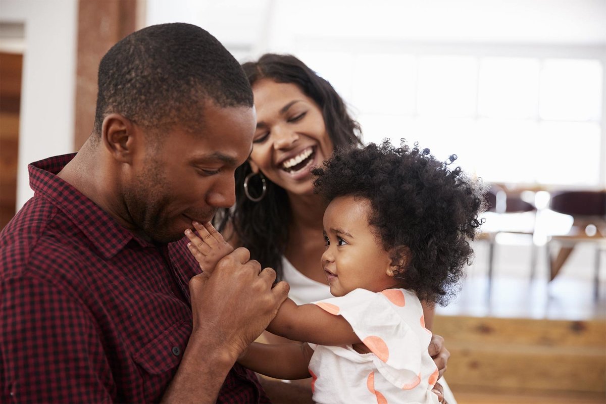 Black father affectionately kissing his toddler's hand, with the mother looking on lovingly, showcasing parental love and care. Join Successful Black Parenting Magazine for more inspiring family moments.