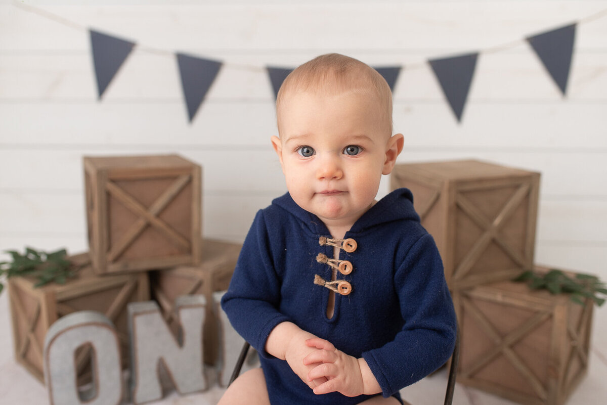 One year old boy smiling at the camera with rustic cake smash set behind him Blue, tan and white cake smash session at studio in Canton, CT |Sharon Leger Photography | Canton, CT Newborn & Family Photographer