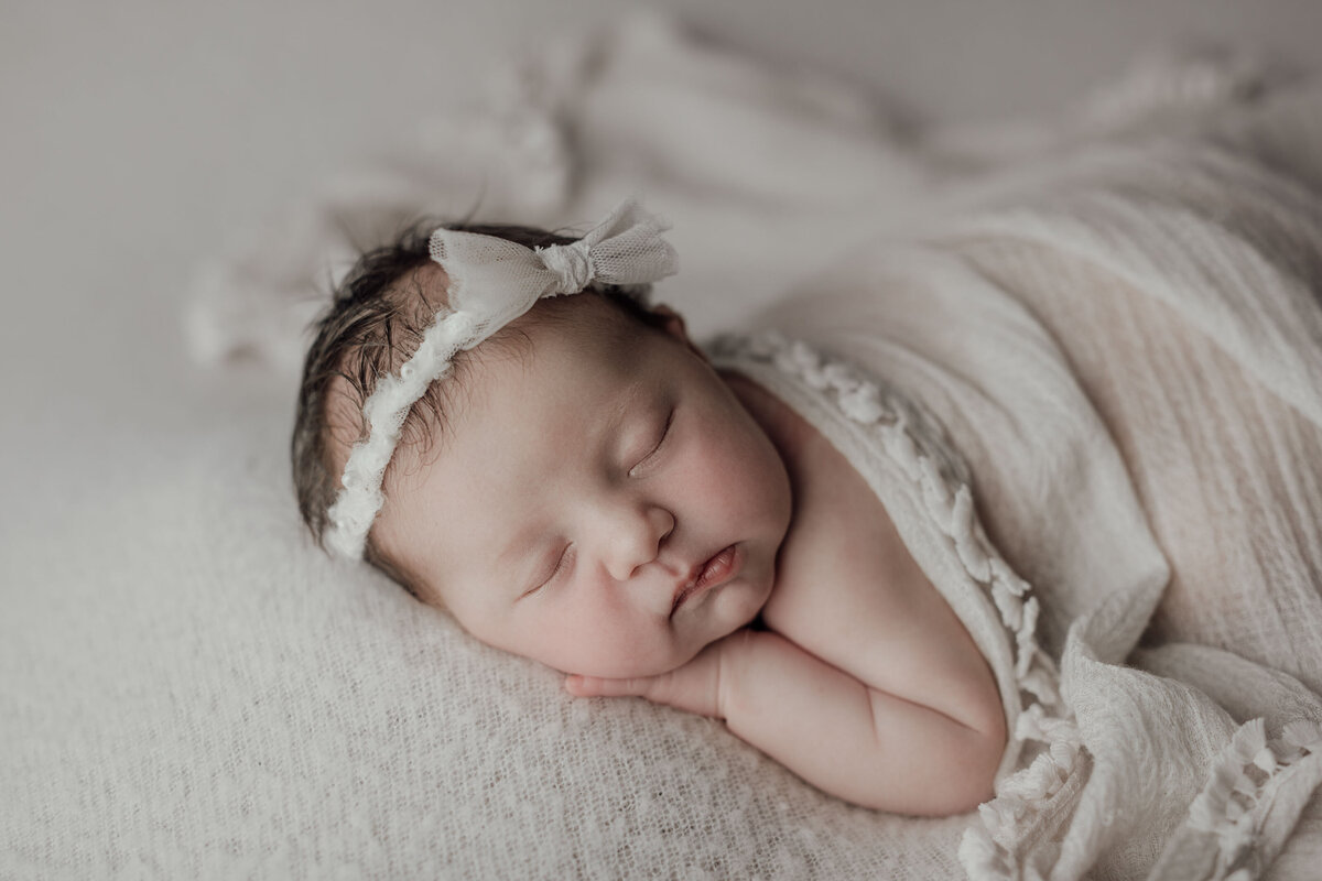 Studio newborn photography - baby sleeping on her belly with her head turned toward the camera. Her hand is resting under her chin. She is loosely covered in a cream blanket and matching cream headband.
