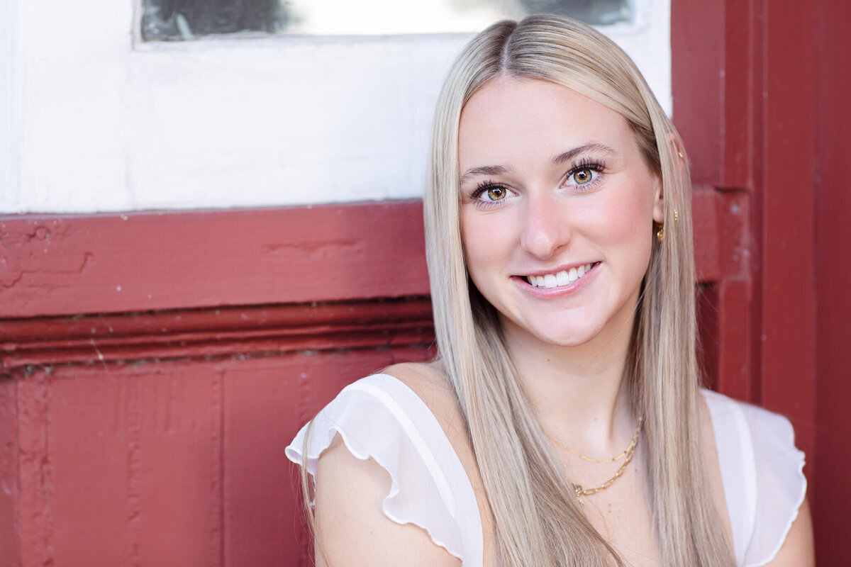 A woman with long blonde hair and a white top smiles in front of a wooden red and white wall.