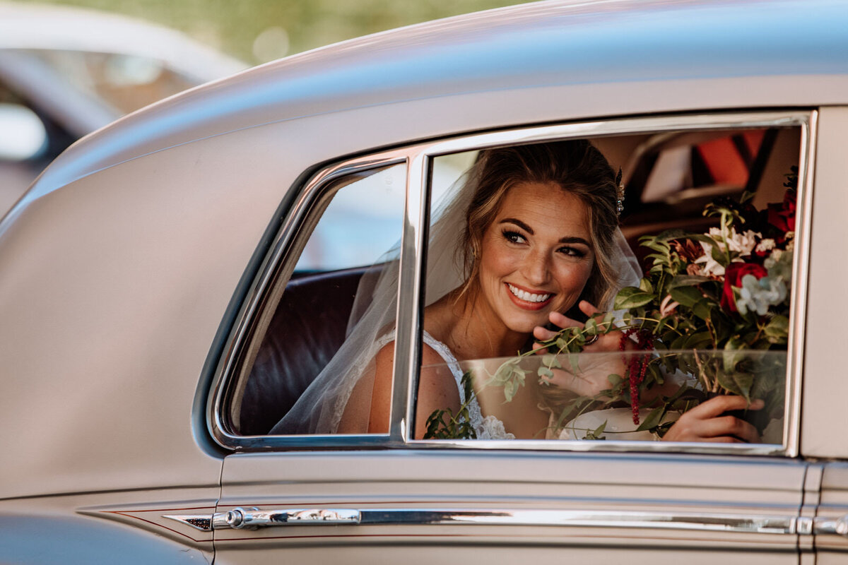 A woman in a bridal gown sitting in a classic car, holding a bouquet of flowers, looking out the car window and smiling and waving