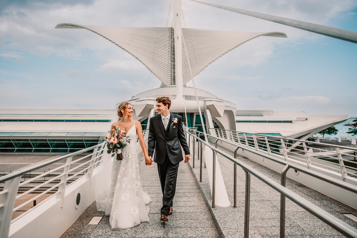A bride and groom walk on a skyway at the Milwaukee Art Museum holding hands and smiling at each other