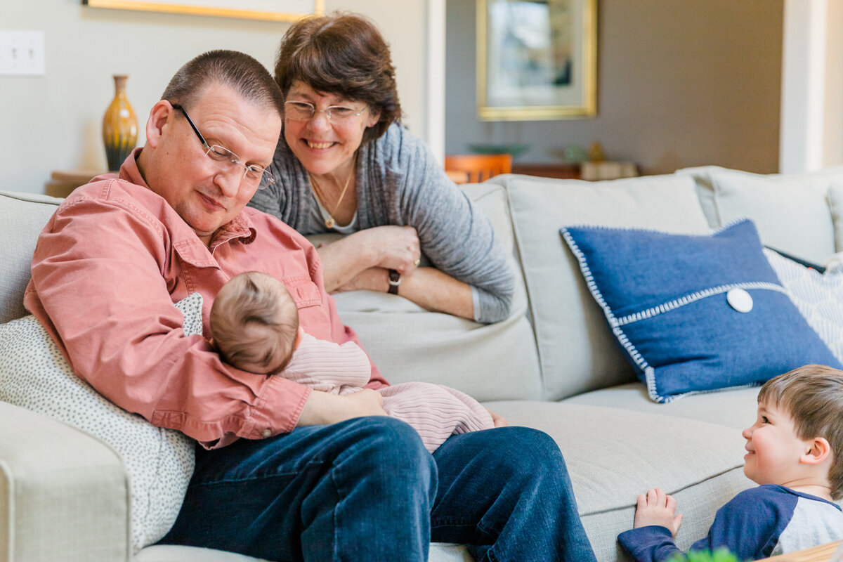 Grandfather sitting on a grey couch smiling at newborn in his arms, his wife smiling down while leaning on the couch, toddler looking on from the floor in the corner