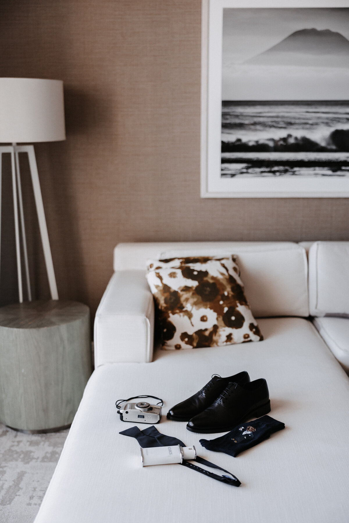 Groom details on a clean white linen couch in hotel room, including shoes, socks, cologne, bowtie and film camera