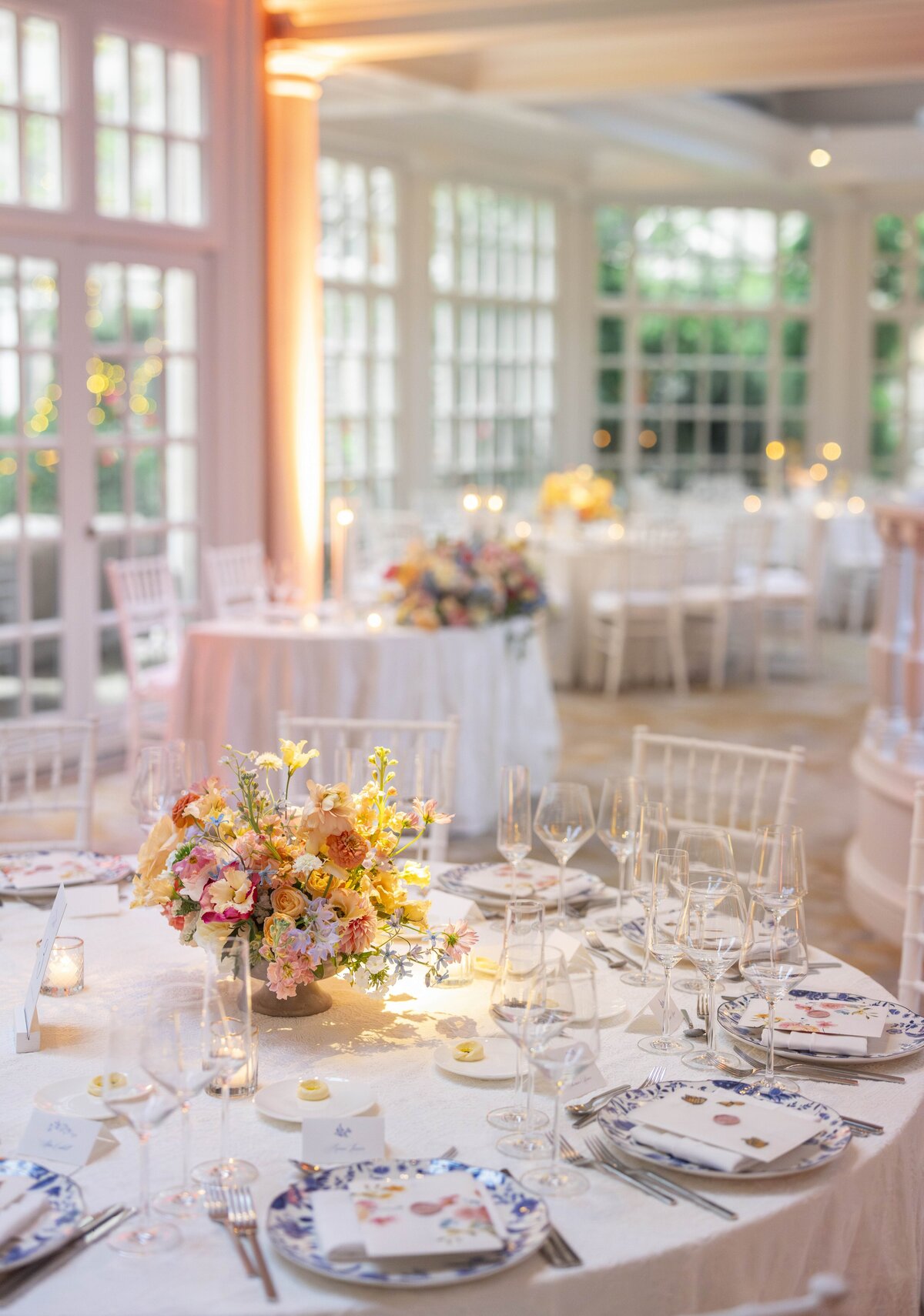 Elegant indoor wedding reception with round tables decorated with floral centerpieces, white tablecloths, and fine china. Soft lighting filters through large windows, creating a warm and inviting atmosphere.