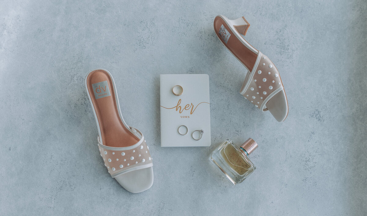 Flat lay of bridal shoes, vow booklet, wedding rings, and perfume bottle on a soft textured surface at Lesner Inn.