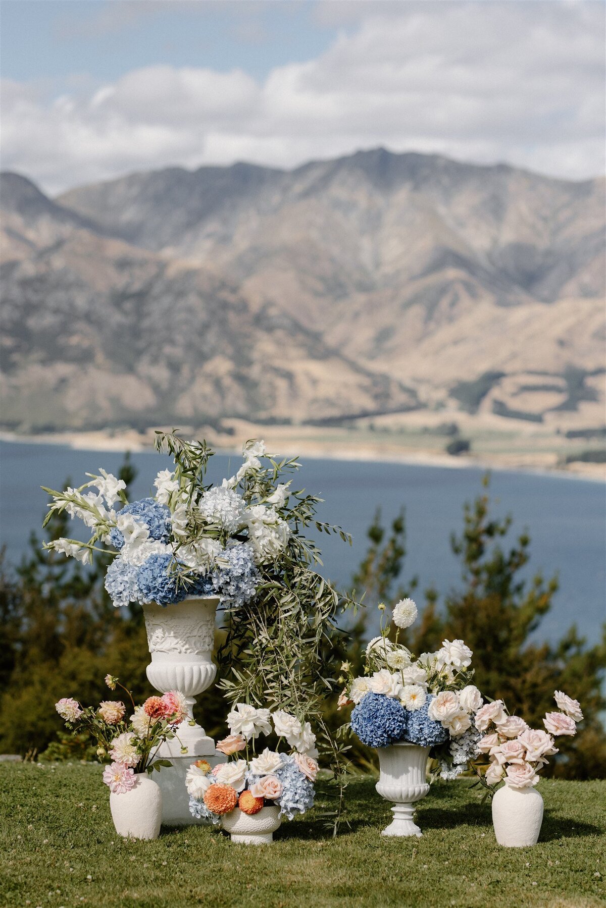 Alfresco wedding ceremony floral setup featuring blue hydrangeas, white gladiolus, and peach dahlias in white urns, overlooking Lake Hāwea.