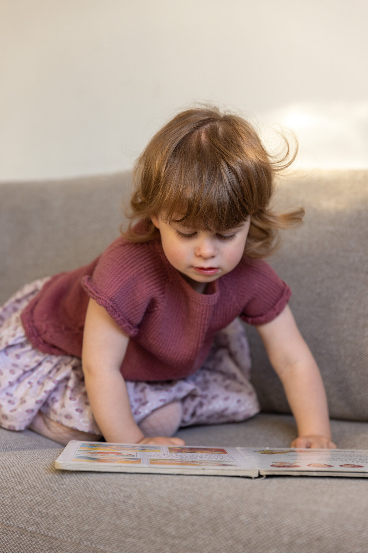 Livsstilsfoto av en jente som sitter på en grå sofa og ser på en bok som ligger på sofasetet.