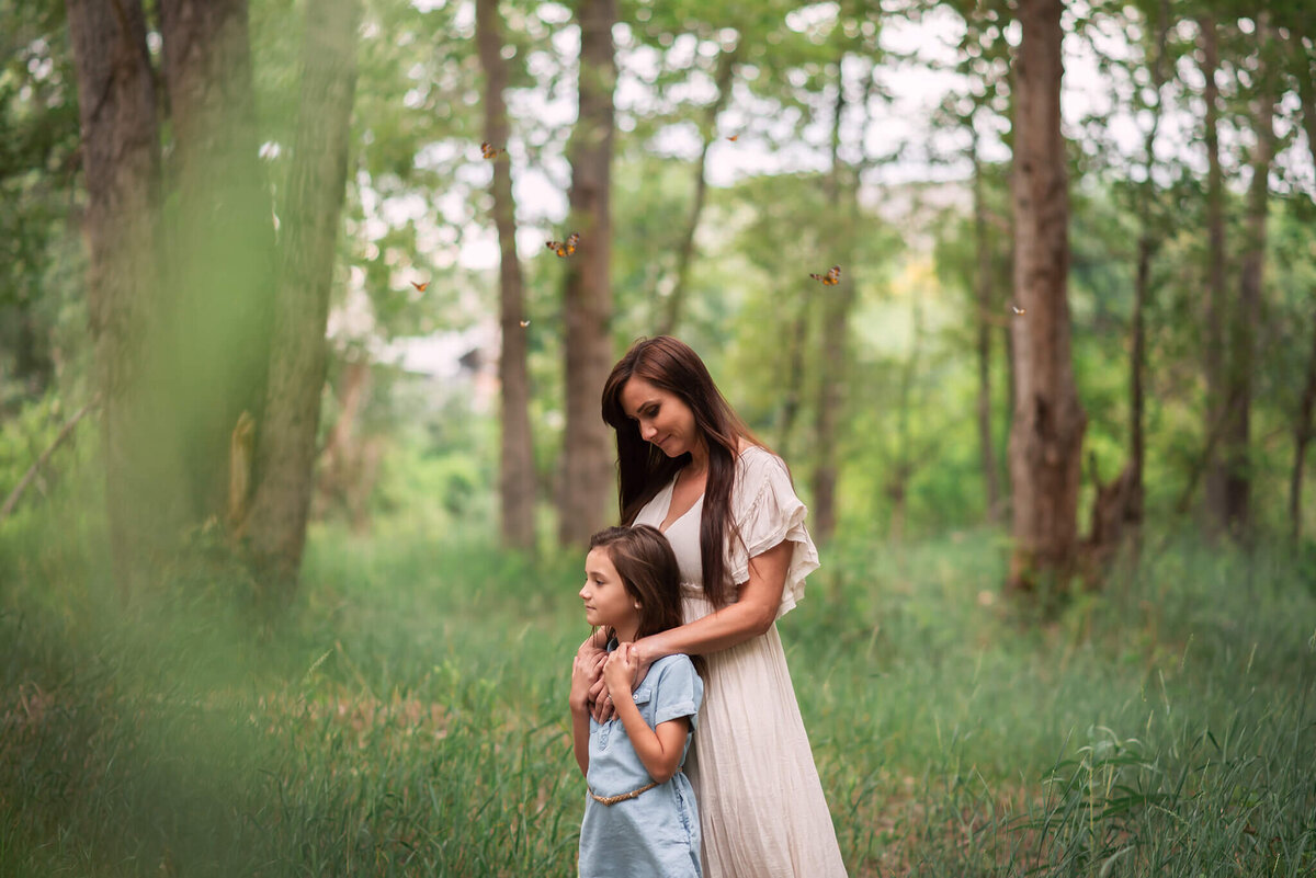A mother embraces her daughter from behind in the tall grass. Butterflies surround them. Taken by Brittany danielle in Kamloops