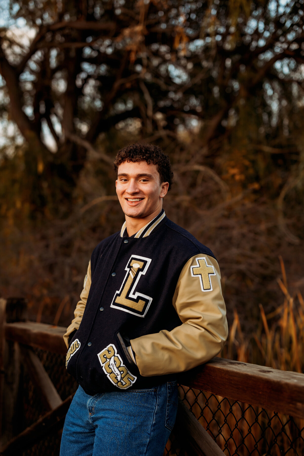 Senior wearing Legacy Lightning Lettermans jacket for senior photos while resting back against wood bridge