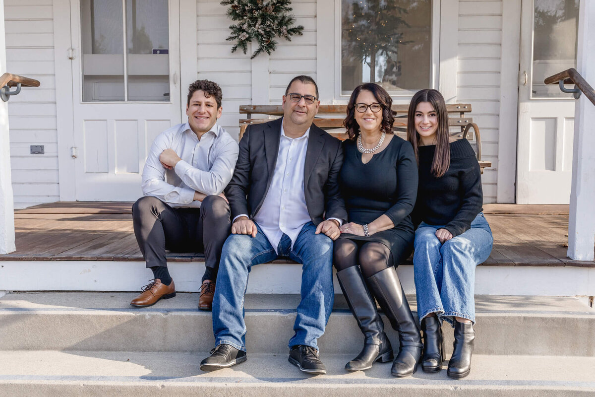 A family of four with brown hair sitting on brown and white porch steps, smiling at the camera.