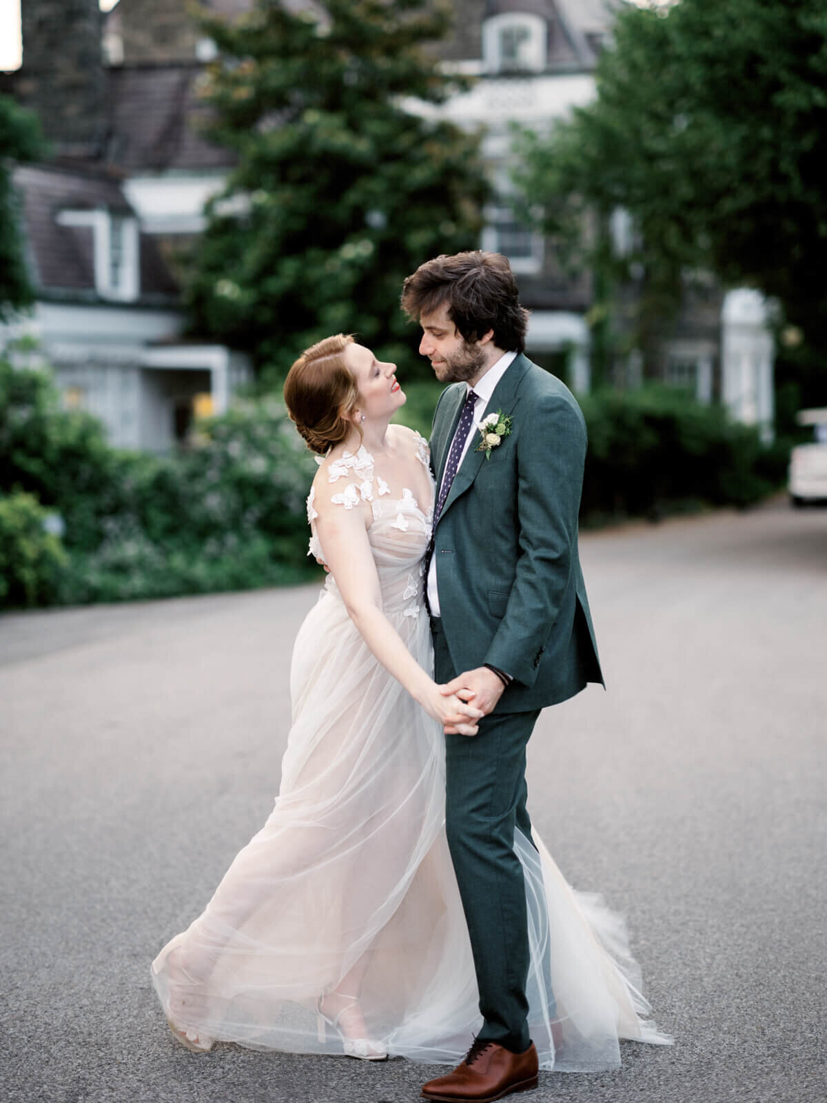 The bride and the groom are standing close together and holding hands, with houses and trees in the background.