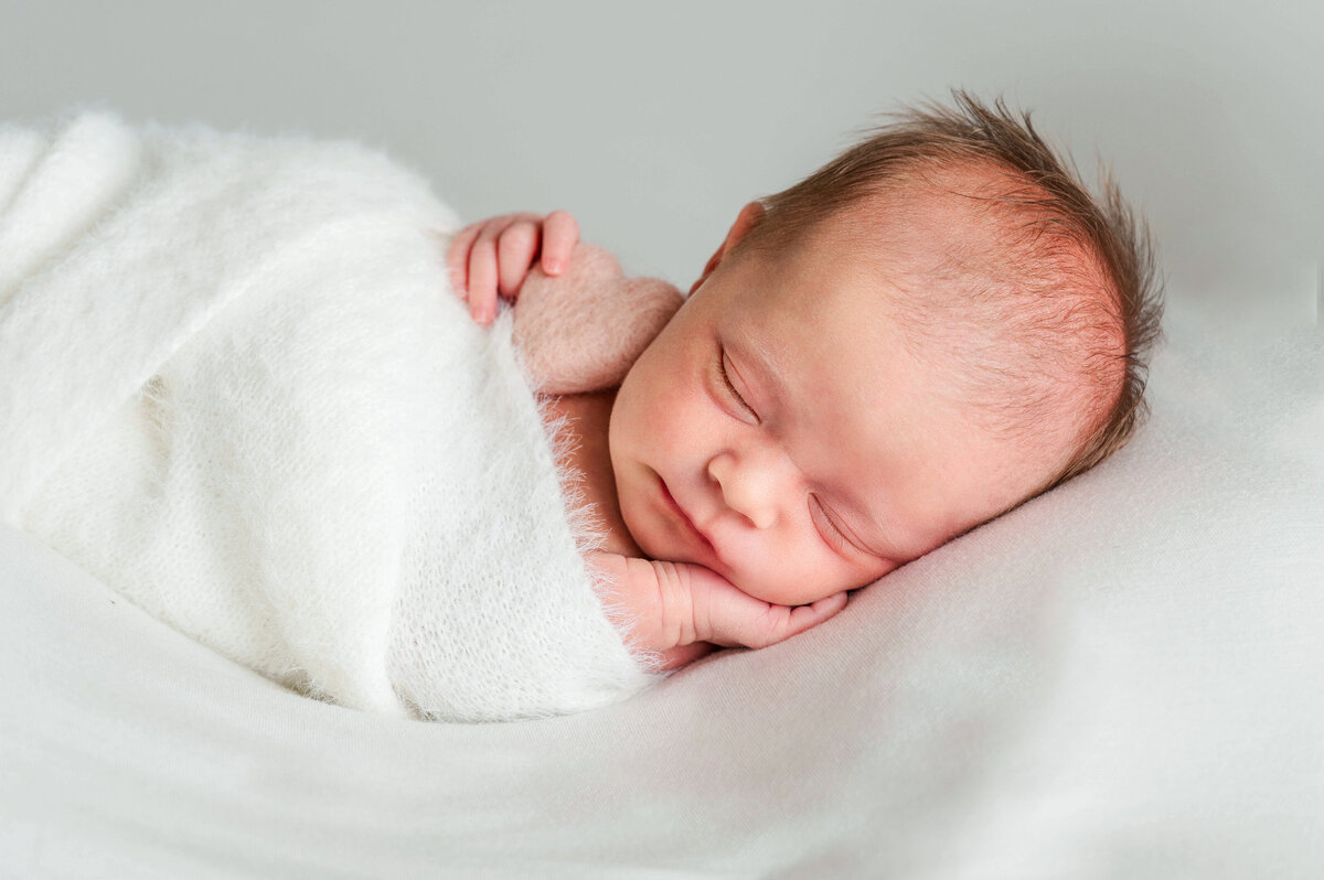Adorable week old baby laying on hand, wrapped in white blanket and holding a small pink heart.