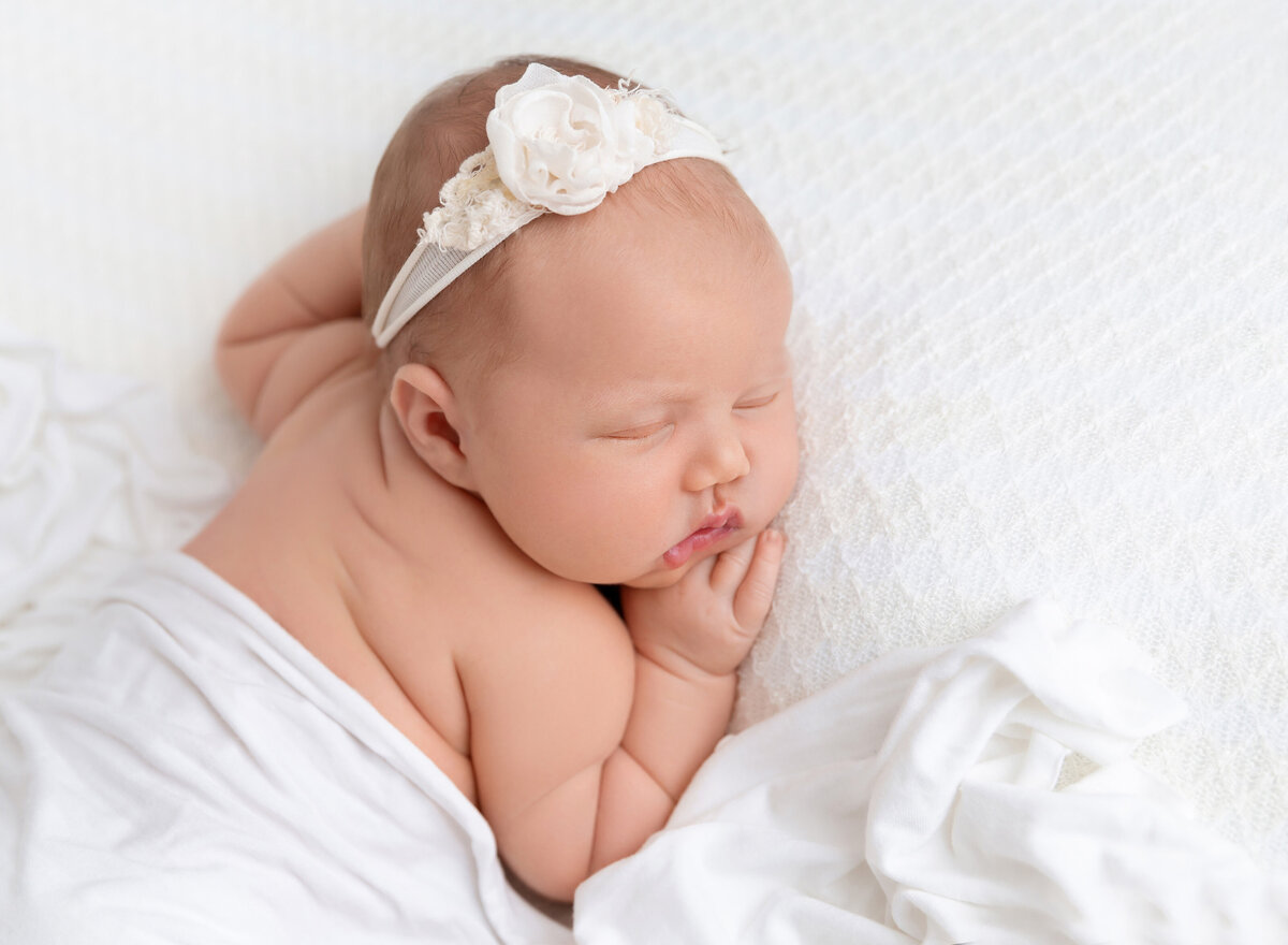 Aerial image. Newborn baby girl photoshoot. Baby is sleeping on her belly with one hand resting under her chin. Baby is draped in white fabric and wearing a white headband.