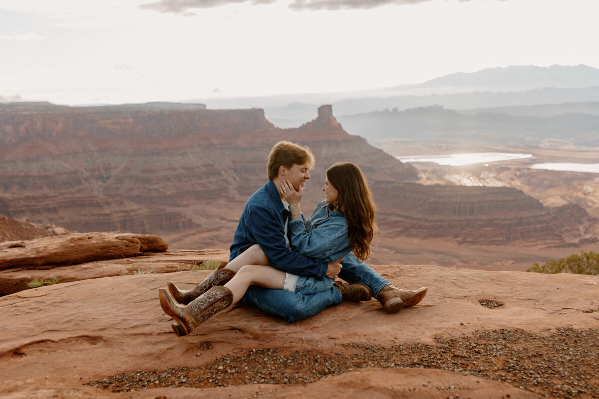 A couple sitting at Dead Horse State Park in Moab. They are both wearing denim, and cowboy boots, and smiling at each other. 