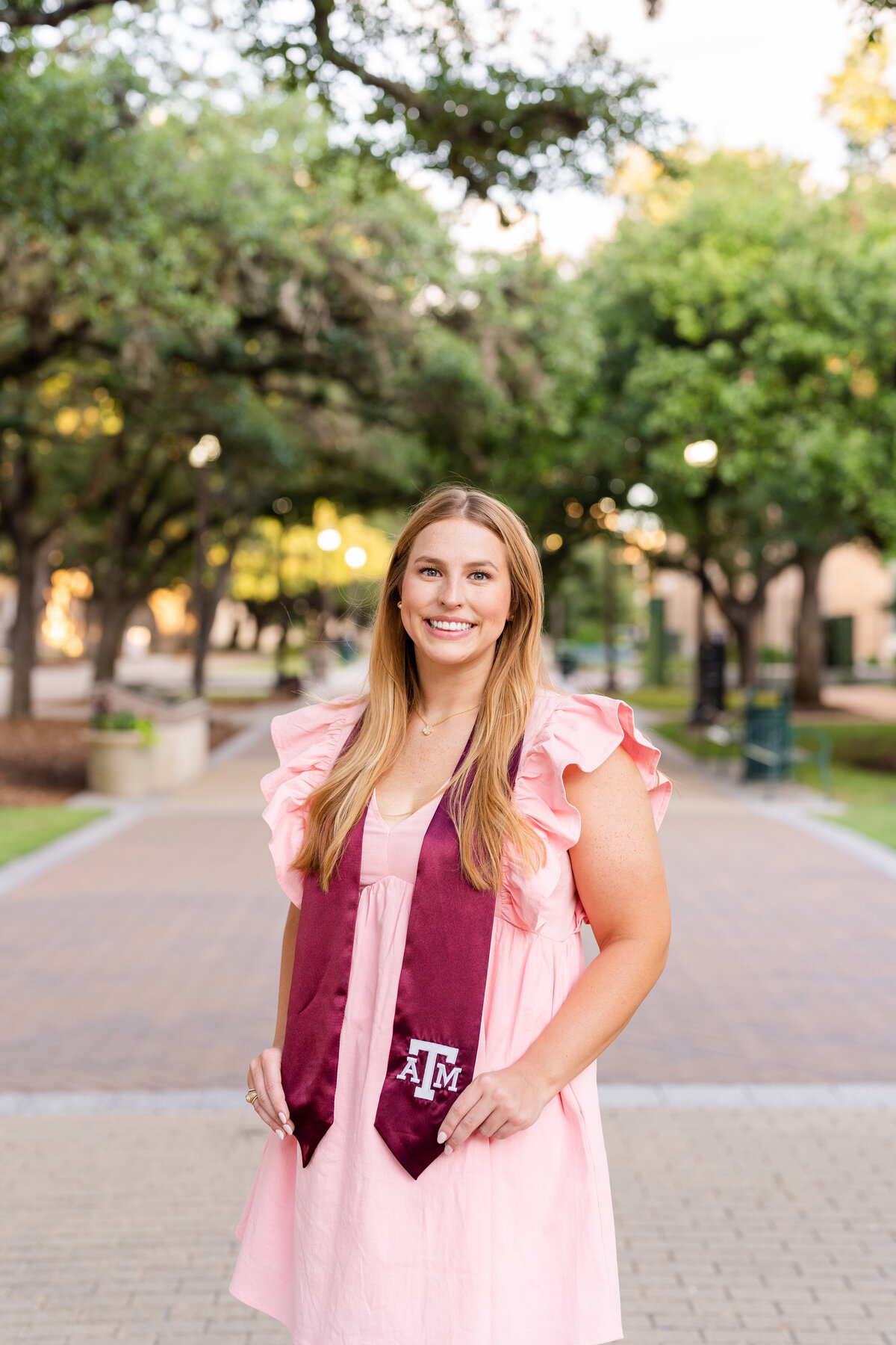 Texas A&M senior girl wearing pink dress and holding Aggie stole and smiling under trees of Military Plaza