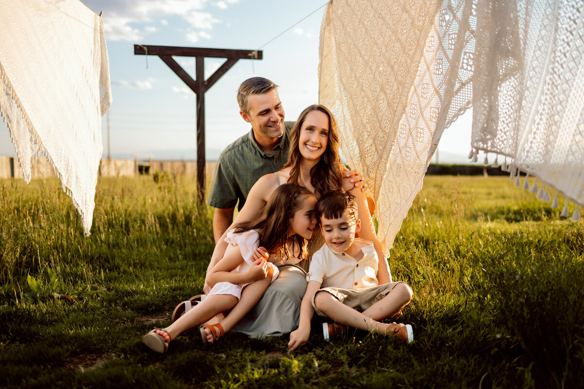 Family of four pile together on farmland on a warm sunny summer day in between lace blankets drying on a clothing line for their Denver family photos
