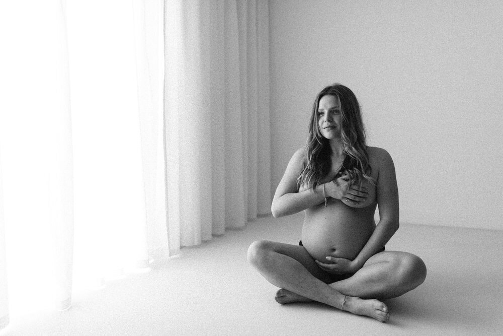 Women sits on the floor crossed legged and smiles, whilst holding her  pregnant belly