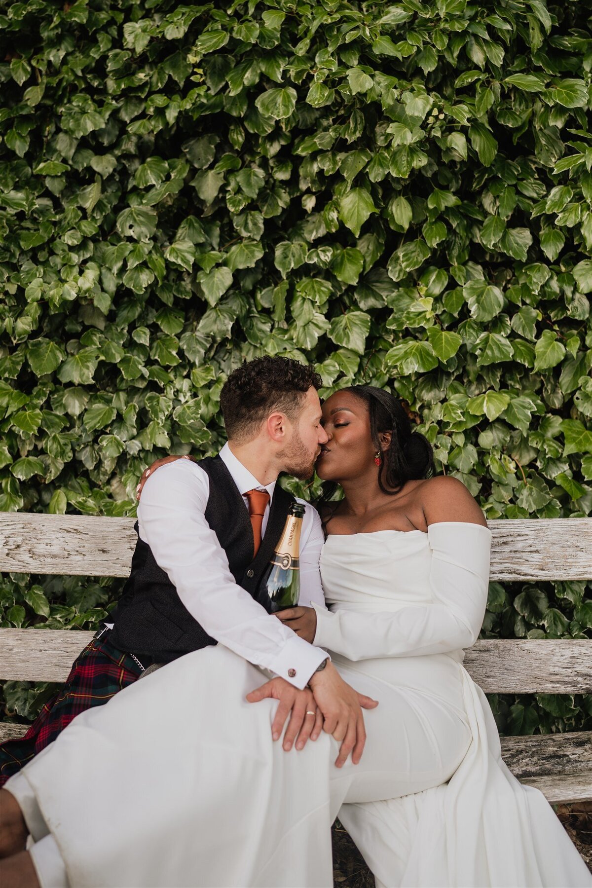 A bride and groom sit on an old bench. The bride rests her legs on the grooms knees as she kisses the groom. The wall behind them is covered in ivy.