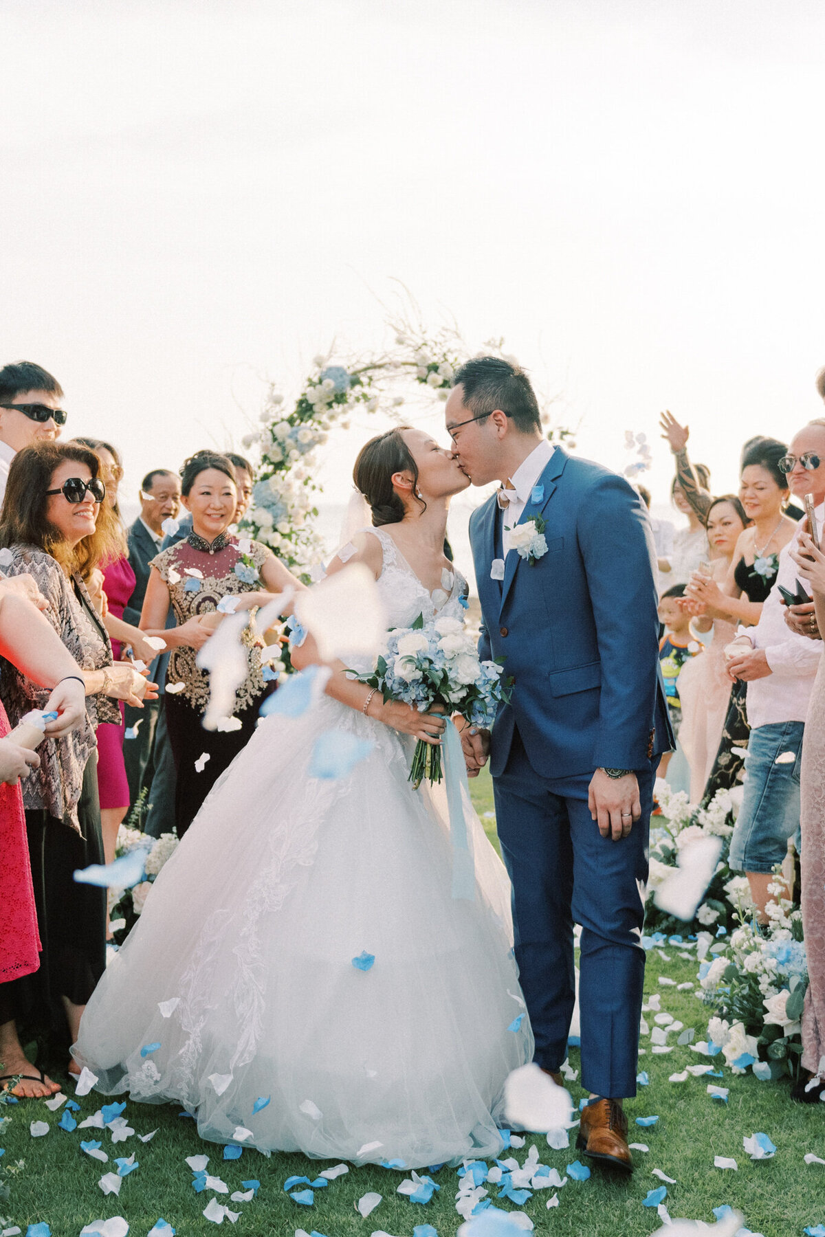 Bride and groom kiss while guests throw blue and white flower petals captured by Pam Kriangkum Photography, fine art, classic wedding photographer in Edmonton, Alberta. Featured on the Bronte Bride Vendor Guide.